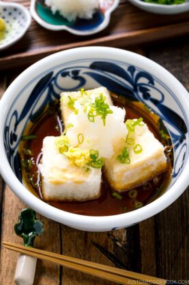 A ceramic bowl containing Agedashi Tofu (Agedashi Dofu) topped with grated daikon, chopped scallions, and grated ginger.