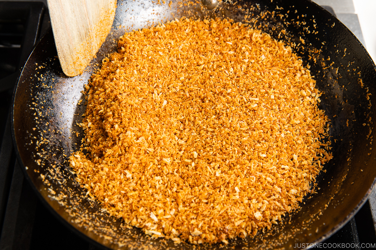 Golden toasted panko breadcrumbs being stirred in a black frying pan with a wooden spatula on a stove.