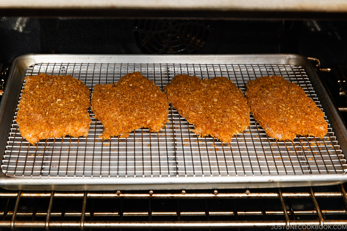 Four breaded chicken cutlets are arranged on a wire rack over a baking sheet inside an oven, being baked.