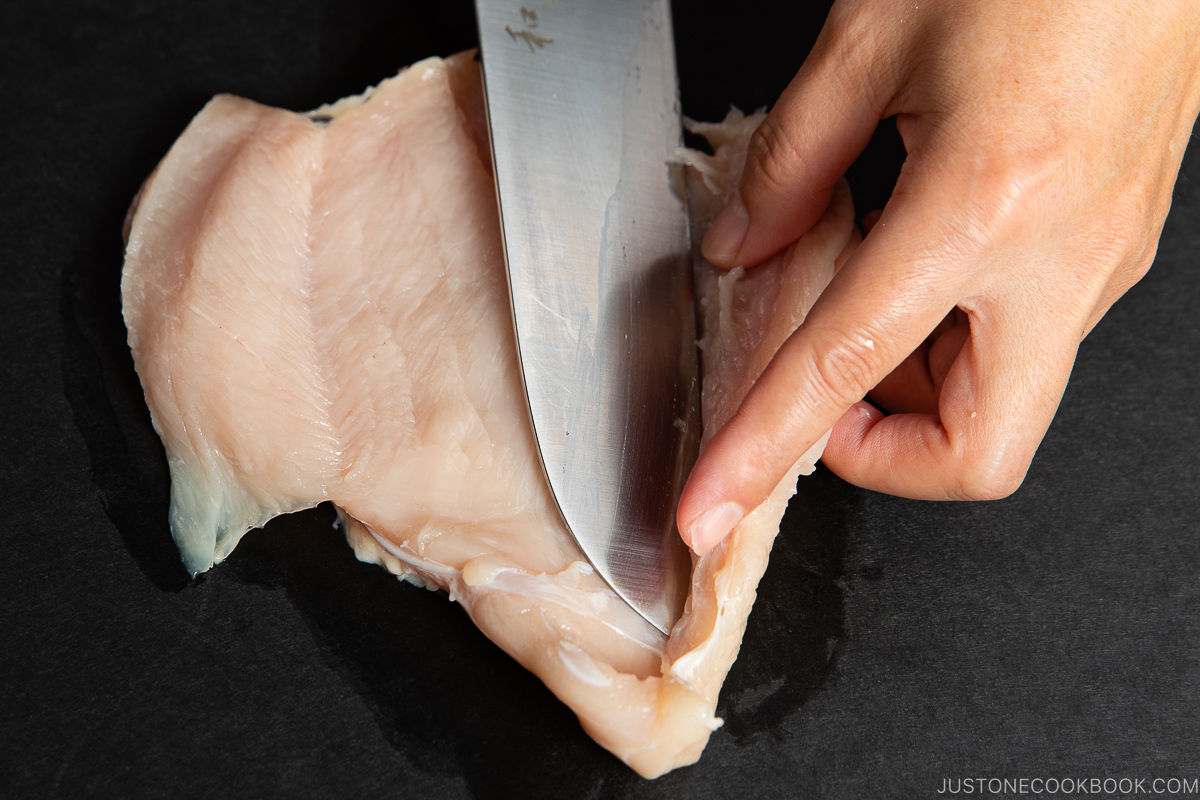 A hand holds a raw chicken breast while using a large knife to slice it in half on a dark surface.