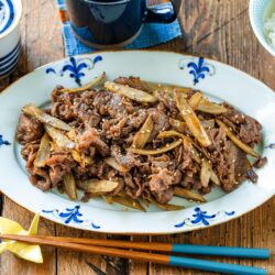 A platter of Beef and Gobo Stir Fry, topped with sesame seeds and served with chopsticks, a bowl of white rice, a cup, and small side dishes on a rustic wooden table.
