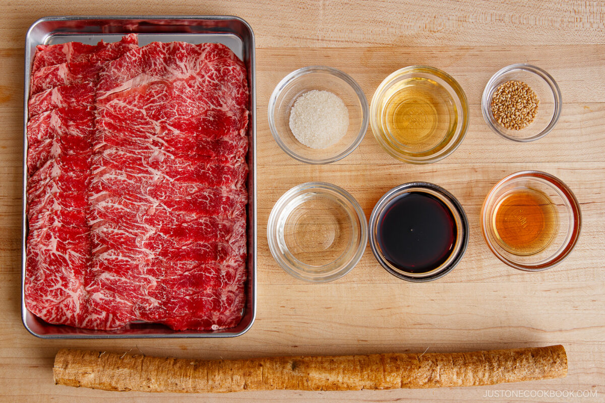 Thinly sliced marbled beef in a tray, six small bowls with various sauces and seasonings, and a long gobo root arranged neatly on a wooden surface—perfect ingredients for making Beef and Gobo Stir Fry.