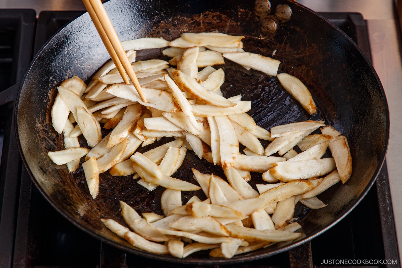 Thin slices of burdock root sizzle in a large black frying pan on the stovetop, stirred with chopsticks for a savory Beef and Gobo Stir Fry.