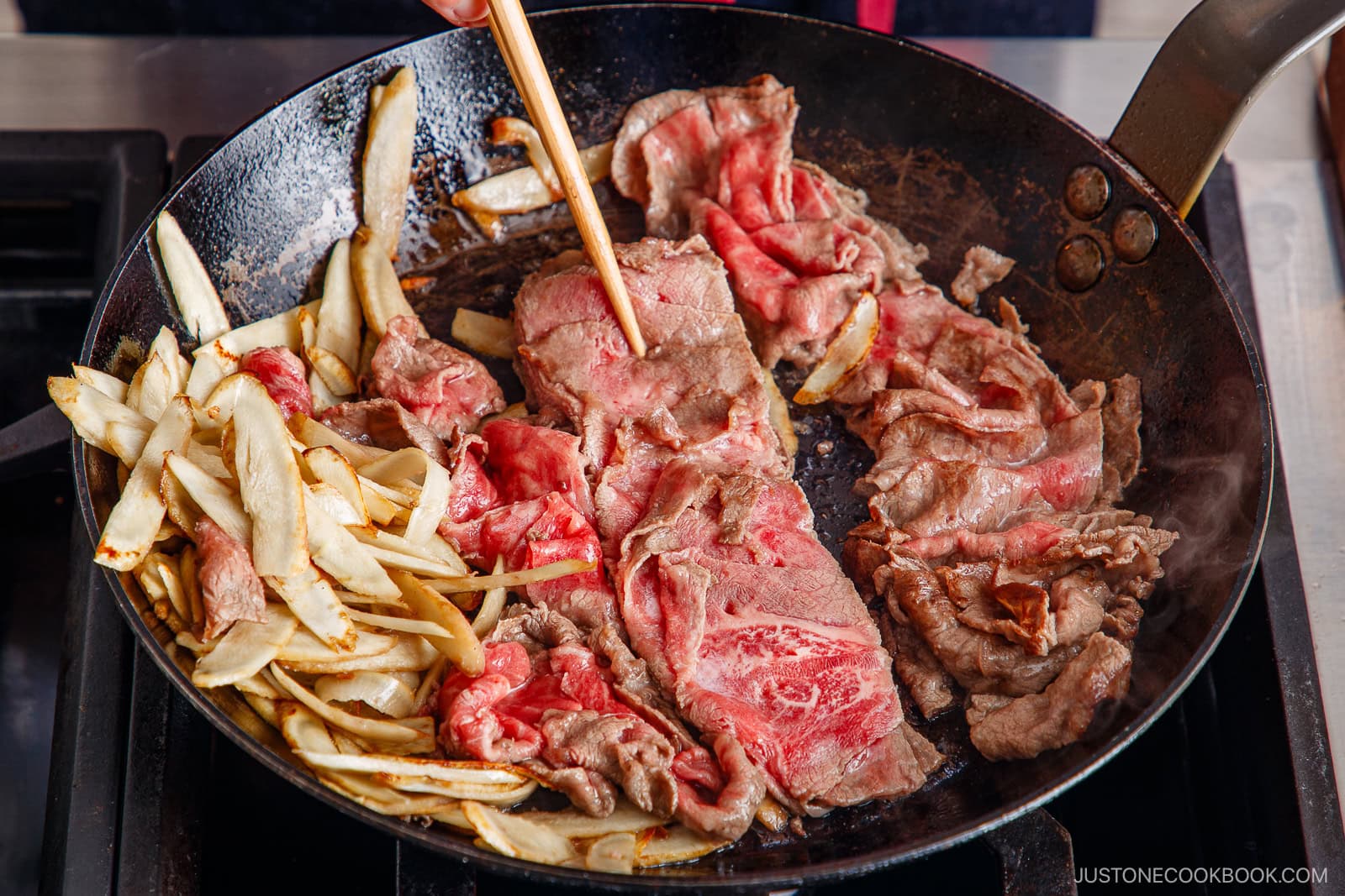 Thinly sliced beef and burdock root are being sautéed for a savory Beef and Gobo Stir Fry in a black skillet. A hand uses chopsticks to cook the beef, which is partially browned and partially raw.