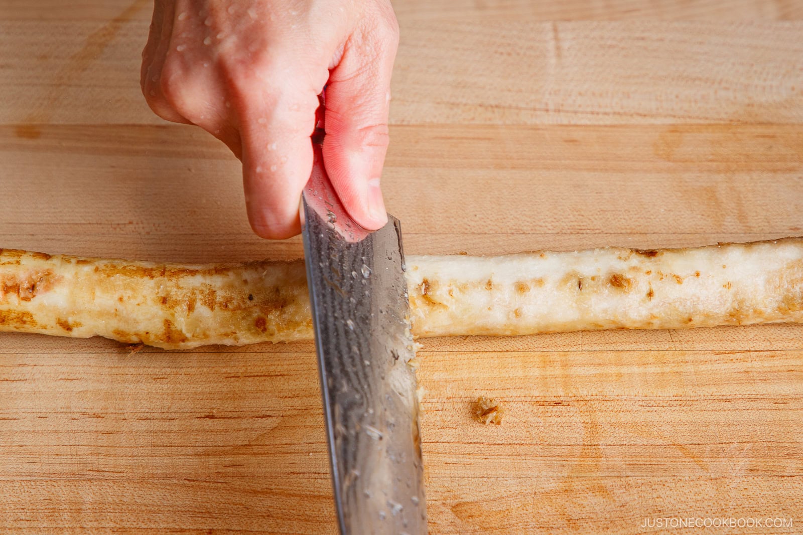A hand uses a knife to cut a rolled cooked food item, possibly an omelette, fish cake, or Beef and Gobo Stir Fry roll, on a wooden cutting board.