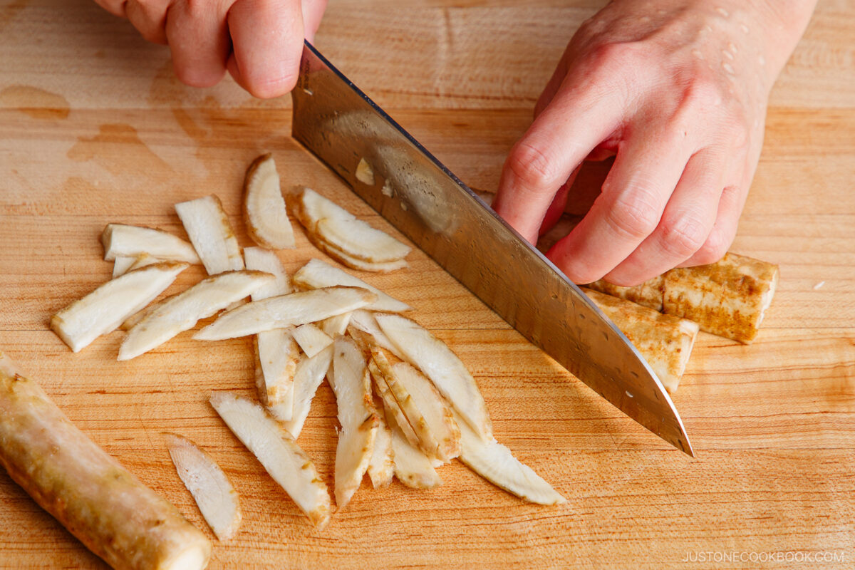 A person slices burdock root into thin strips on a wooden cutting board with a large kitchen knife, preparing ingredients for a delicious Beef and Gobo Stir Fry.