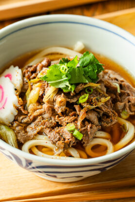A bowl of beef udon with sliced beef, green onions, narutomaki (fish cake), and fresh herbs in broth, served on a wooden tray with chopped green onions and a bottle of seasoning on the side.