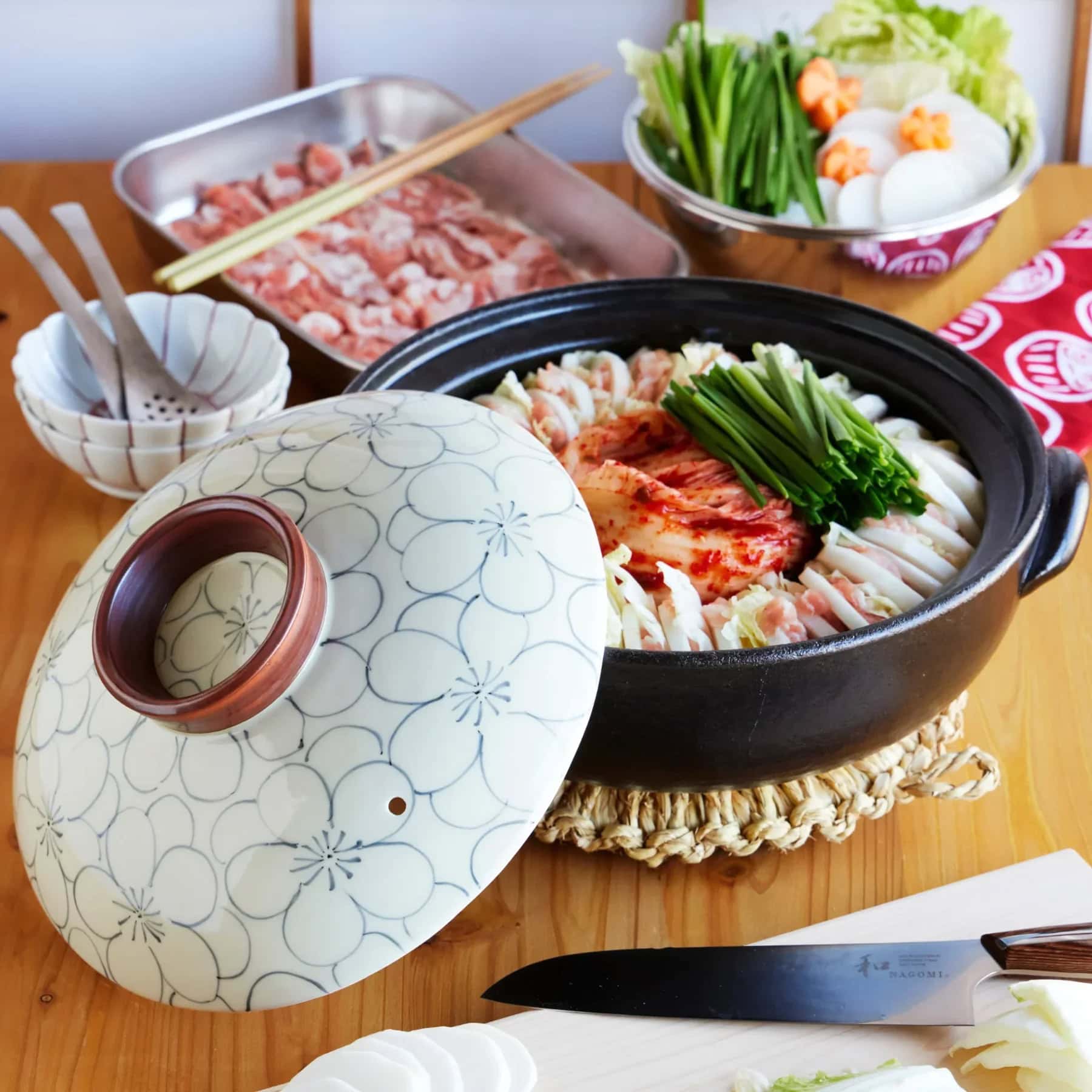 A pot of nabe, or Japanese hot pot, with napa cabbage, sliced meat, greens, and kimchi sits on a wooden table. Nearby are raw meat slices, vegetables, eggs, bowls, and a knife on a cutting board.
