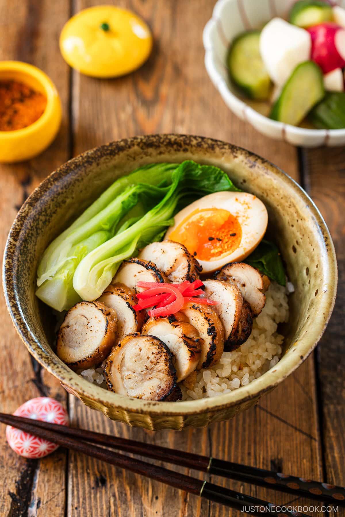 A donburi bowl containing steamed rice topped with seared sliced chicken chashu, soft boiled egg, and blanched bok choy, garnished with pickled ginger.