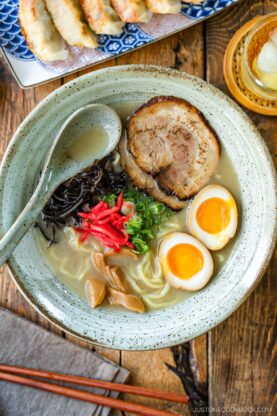 A rustic bowl containing noodles with tonkotsu broth, topped with chashu, ramen eggs, menma, kikurage, ginger, and chopped green onion. On the table, a plate of crispy gyoza and a glass of mugicha are served together.