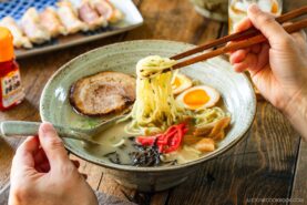 A rustic bowl containing noodles with tonkotsu broth, topped with chashu, ramen eggs, menma, kikurage, ginger, and chopped green onion. On the table, a plate of crispy gyoza and a glass of mugicha are served together.