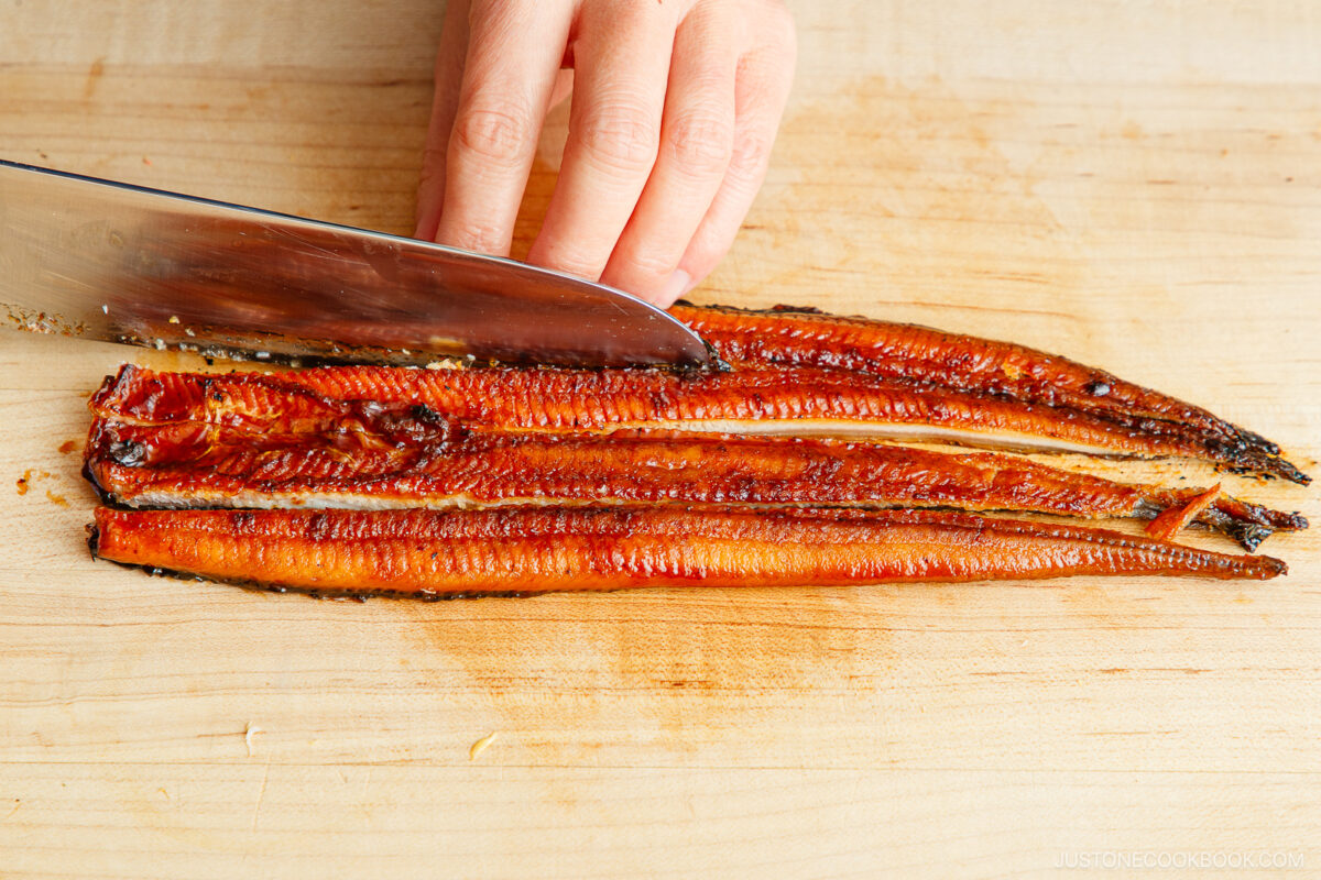 A hand uses a knife to slice a grilled eel fillet on a wooden cutting board, preparing it as a savory filling for ehomaki (setsubun sushi roll).