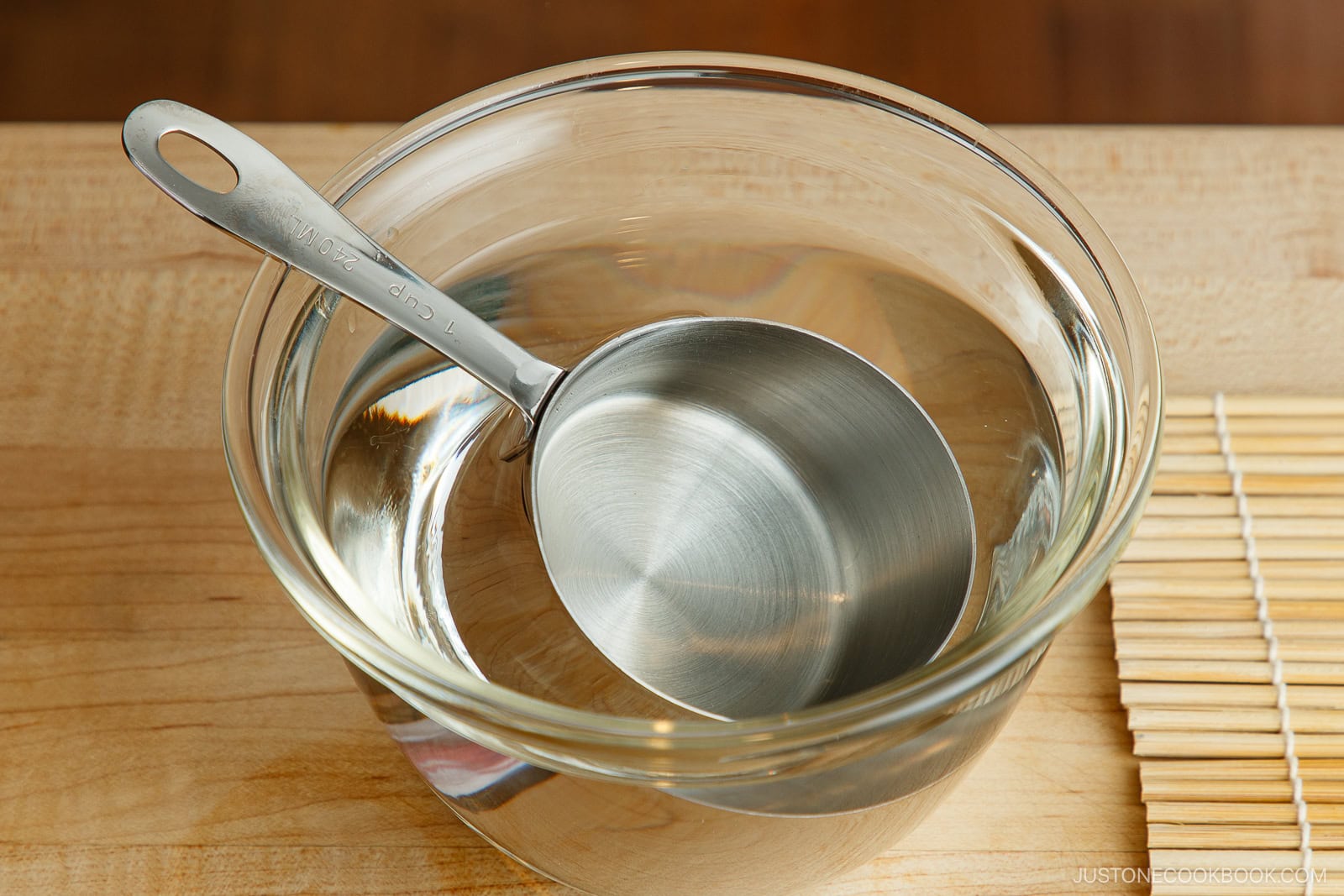 A metal measuring cup filled with water rests inside a clear glass bowl of water on a wooden surface, beside a bamboo mat—perfect for preparing ehomaki (setsubun sushi roll).
