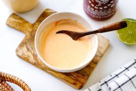 A bowl of creamy orange sauce—your homemade spicy mayo recipe—sits with a wooden spoon on a cutting board. Nearby are a halved lime, sriracha bottle, and checkered cloth on a white surface.
