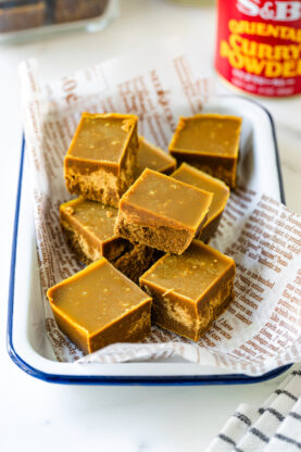 A tray lined with printed paper holds several neatly cut squares of golden brown curry fudge. A can of curry powder sits blurred in the background.