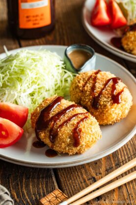 A round white plate containing Japanese croquettes, shredded cabbage, tomato wedges, and a tiny container with sesame dressing.