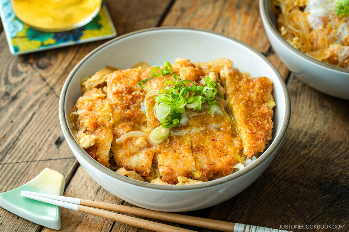 A white bowl containing Katsudon, pork cutlet rice bowl, where golden brown tonkatsu, drizzled egg, and tender onion are simmered in a savory dashi broth.
