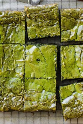 A batch of matcha brownies sliced into nine squares sits on parchment paper, with a small dish of green matcha powder and a purple-patterned napkin nearby.