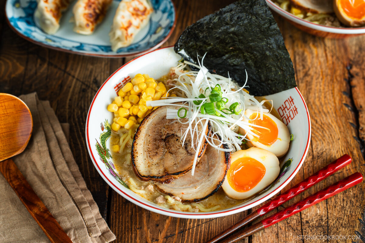 A bowl of ramen with sliced pork, soft-boiled eggs, corn, green onions, seaweed, and noodles in broth. Red chopsticks and a wooden spoon are nearby. Gyoza dumplings are in the background on a wooden table.
