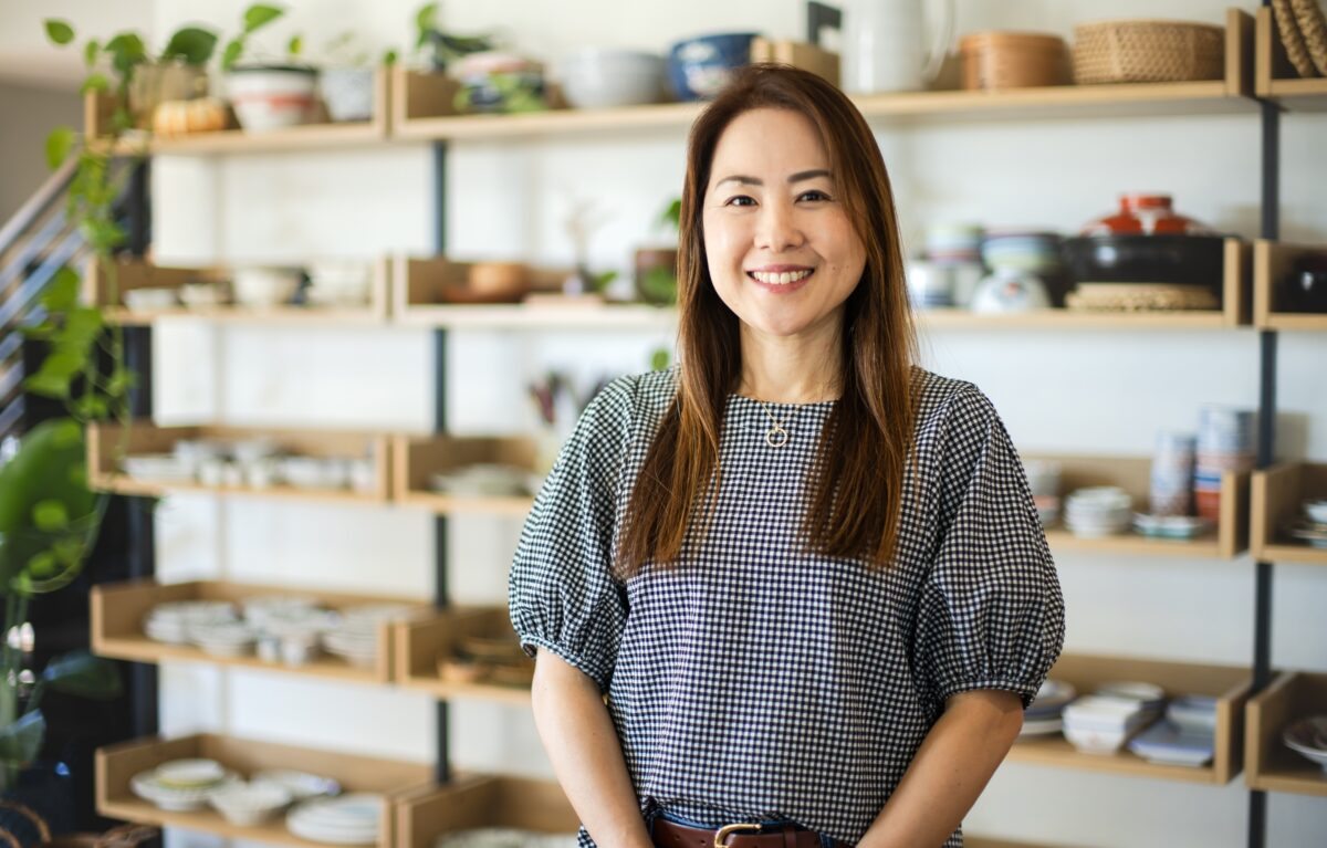 A woman with long hair, wearing a checkered blouse, stands smiling in front of shelves filled with various dishes and pottery in a well-lit room—perfectly capturing the warm atmosphere of About Just One Cookbook.
