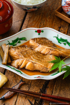 Two pieces of simmered fish fillet with a light brown glaze, served on a decorative plate with sauce, burdock root, and a green maple leaf, set on a wooden table with rice and miso soup in the background.