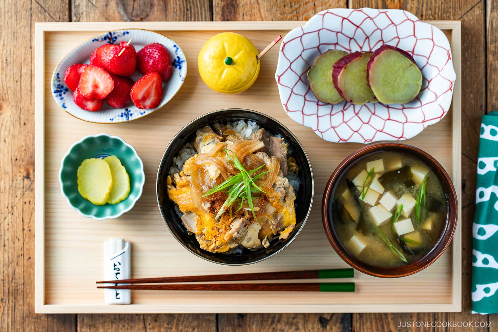 A Japanese meal set on a wooden tray includes a bowl of rice with meat and scallions, miso soup with tofu, sliced strawberries, sweet potato slices, pickles, chopsticks, and a small yellow condiment container.