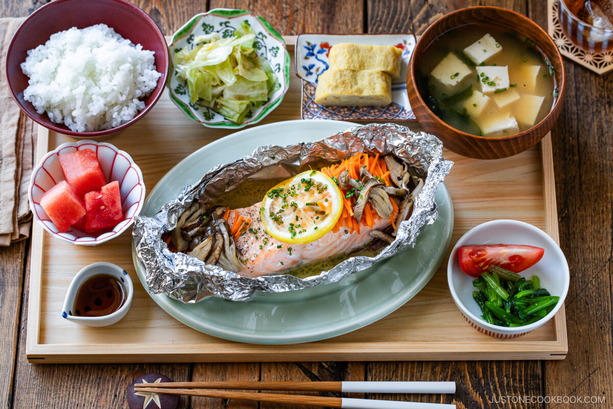 A Japanese meal set with grilled salmon in foil with lemon and vegetables, rice, miso soup with tofu, pickled greens, tamagoyaki, cabbage, watermelon, tomato, and soy sauce, all served on a wooden tray.