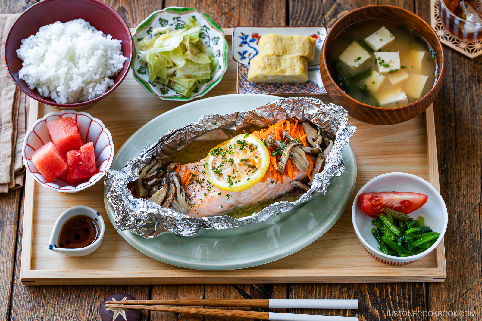 A Japanese meal set with grilled salmon in foil with lemon and vegetables, rice, miso soup with tofu, pickled greens, tamagoyaki, cabbage, watermelon, tomato, and soy sauce, all served on a wooden tray.