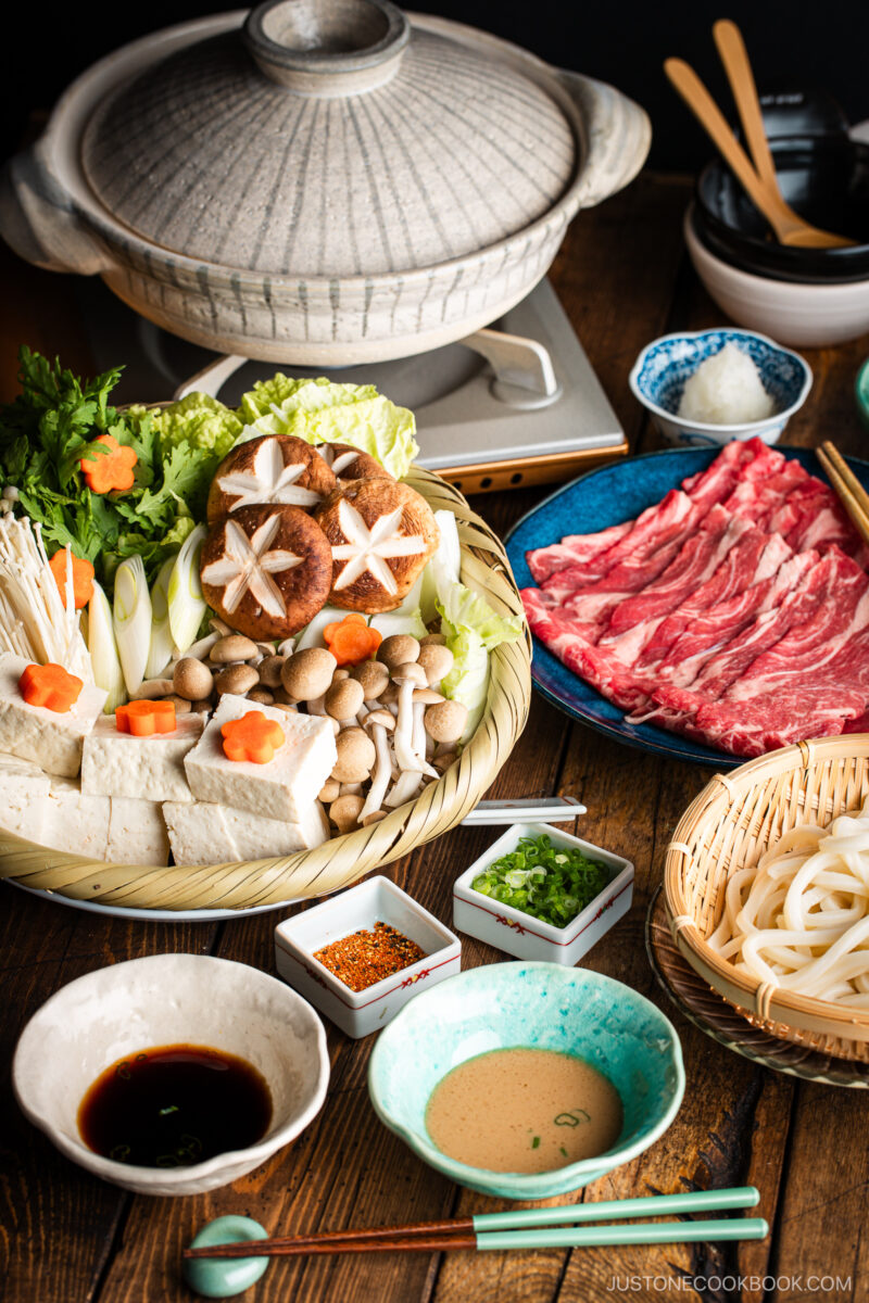 A Japanese shabu-shabu hot pot setup with a donabe pot, thinly sliced beef, tofu, assorted mushrooms, vegetables, udon noodles, and dipping sauces arranged on a wooden table.