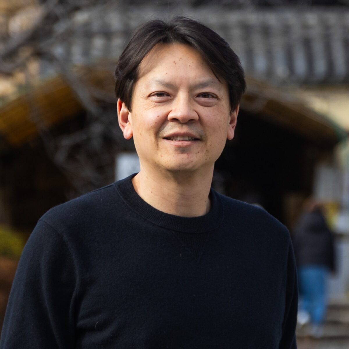Photograph of a smiling man outdoors, wearing a black sweater, with traditional Japanese architecture in the background, representing Japanese culture and cuisine.