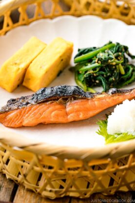 A white plate containing Japanese salted salmon (Shiozake), tamagoyaki, and spinach salad.
