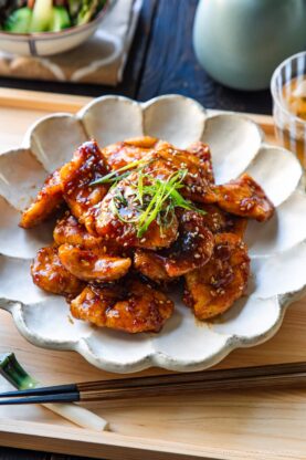 A plate of glazed, pan-fried chicken pieces garnished with thinly sliced green onions and sesame seeds, served on a ceramic dish with chopsticks on a wooden tray.