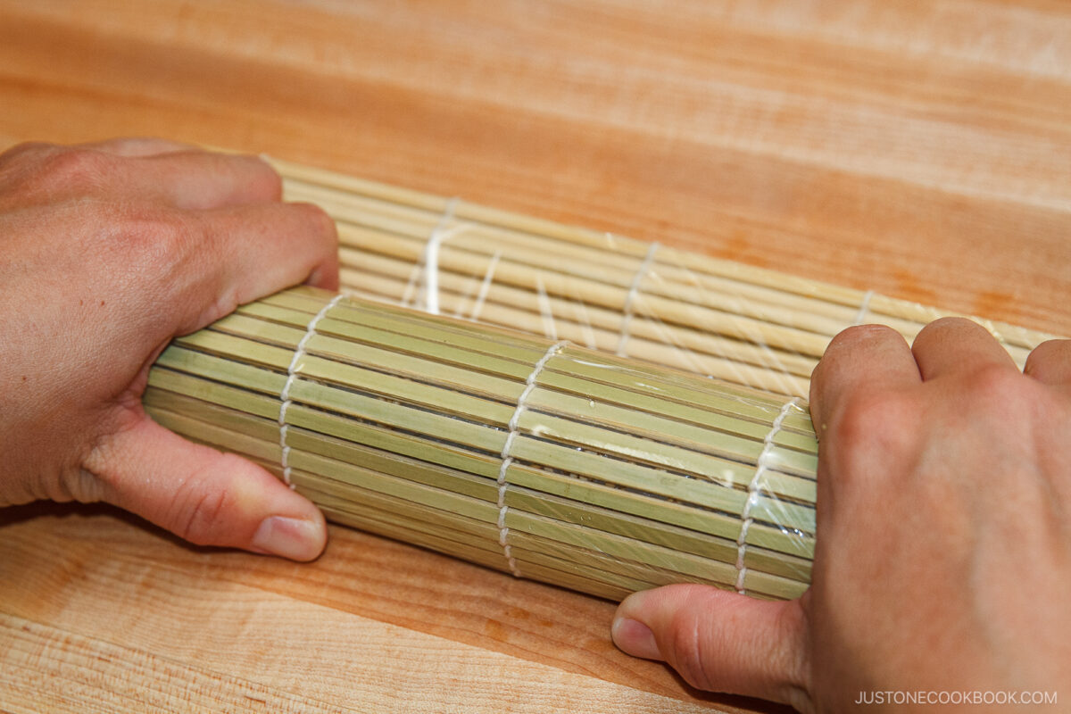 Close-up of hands rolling a bamboo sushi mat on a wooden surface, demonstrating the process of making spicy tuna rolls.