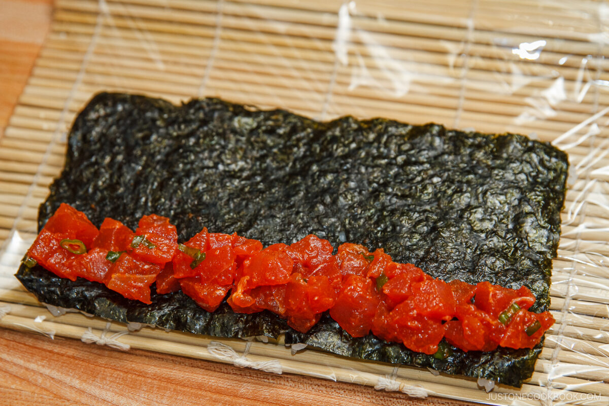 A sheet of nori seaweed on a bamboo sushi mat, topped with a row of diced red tuna, ready to be rolled into spicy tuna rolls or fashioned into a delicious hand roll.