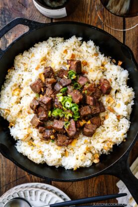 A cast-iron pan containing Steak Garlic Rice topped with chopped scallions and garlic chips.