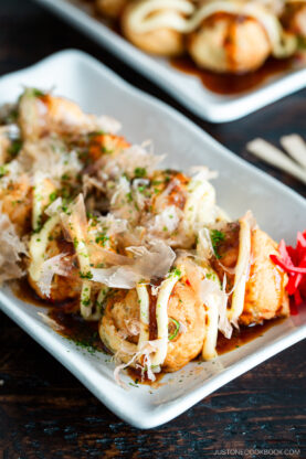 A rectangular white plate with several golden-brown takoyaki balls topped with mayonnaise, sauce, green seaweed flakes, and bonito flakes, garnished with pickled ginger on the side.