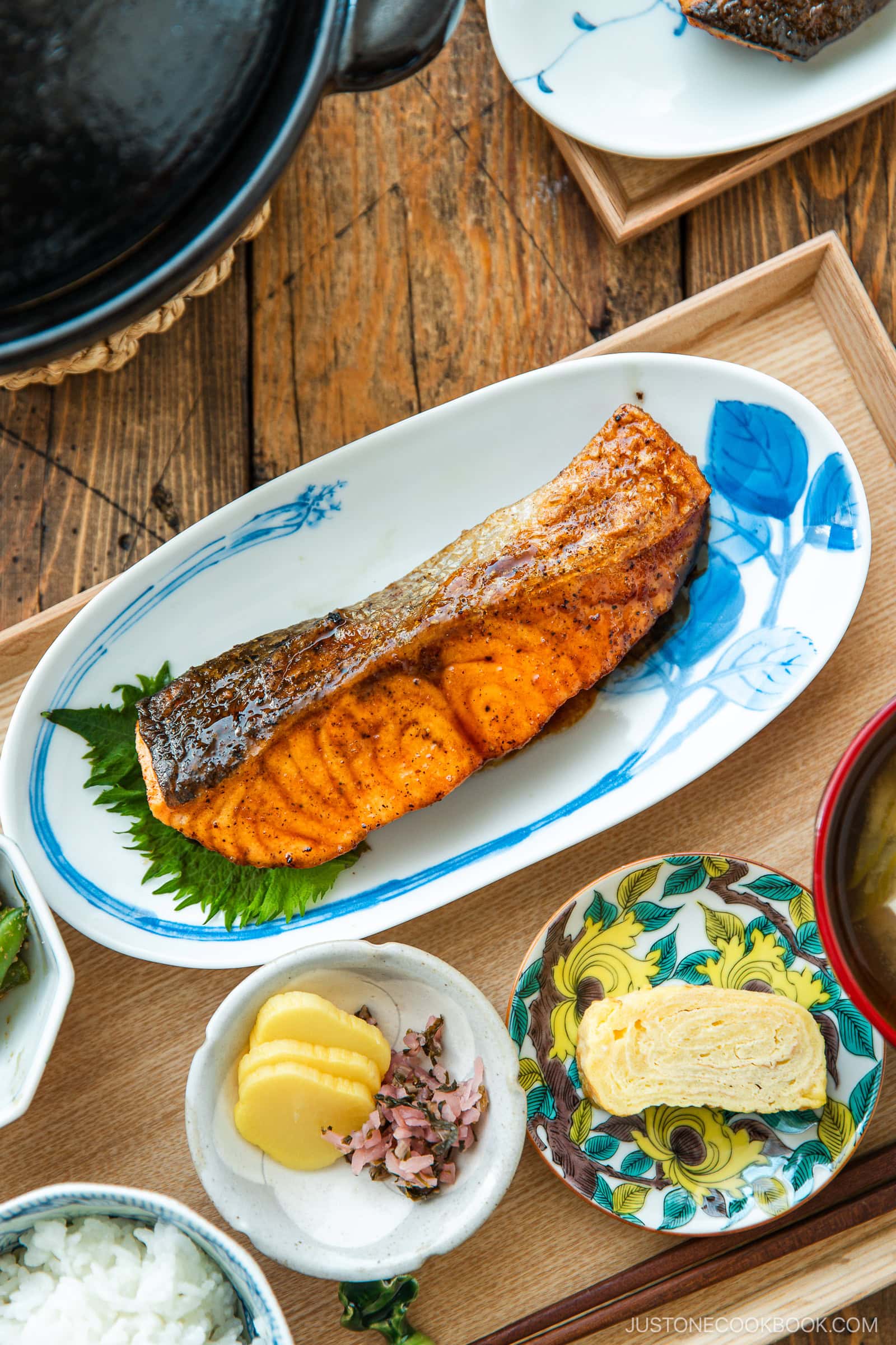 A Japanese meal consisting of small dishes or bowls, each plating Teriyaki Salmon, steamed rice, miso soup, pickles, tamagoyaki, and green bean gomaae.