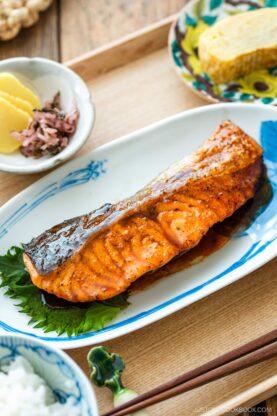 A Japanese meal consisting of small dishes or bowls, each plating Teriyaki Salmon, steamed rice, miso soup, pickles, tamagoyaki, and green bean gomaae.