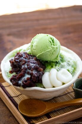 A bowl containing shaved ice, drizzled with matcha syrup and topped with red bean paste, shiratama dango, sweetened condensed milk, and green tea ice cream.