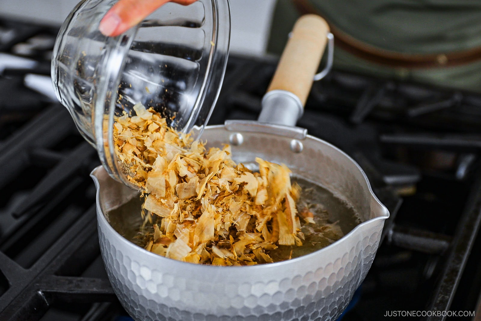 A hand pours dried bonito flakes from a glass bowl into a pot of simmering water on a stove, preparing Japanese dashi broth.