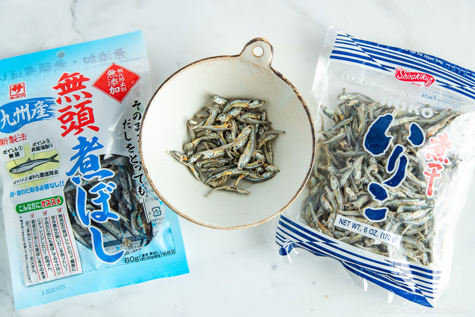 A bowl of dried small fish sits between two packages of dried anchovies on a white surface. One package has blue and white Japanese text, the other is clear with blue writing and shows the fish inside.