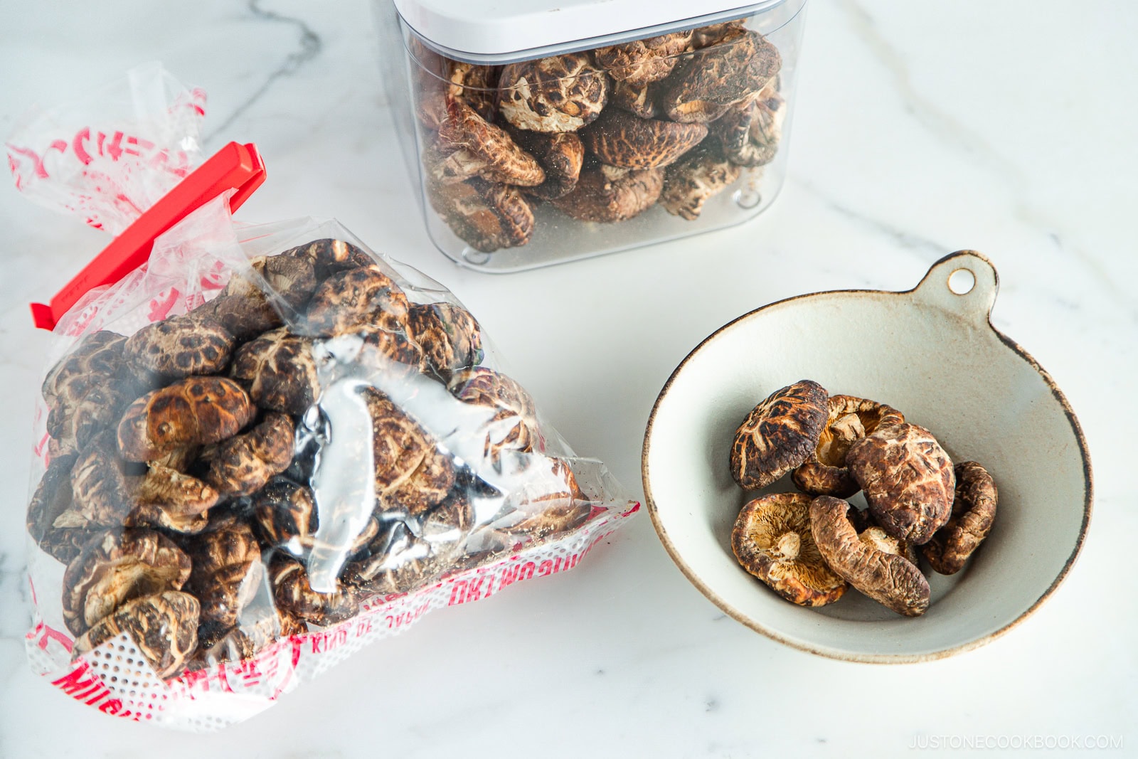 A bag, a container, and a bowl filled with dried shiitake mushrooms are displayed on a white surface.