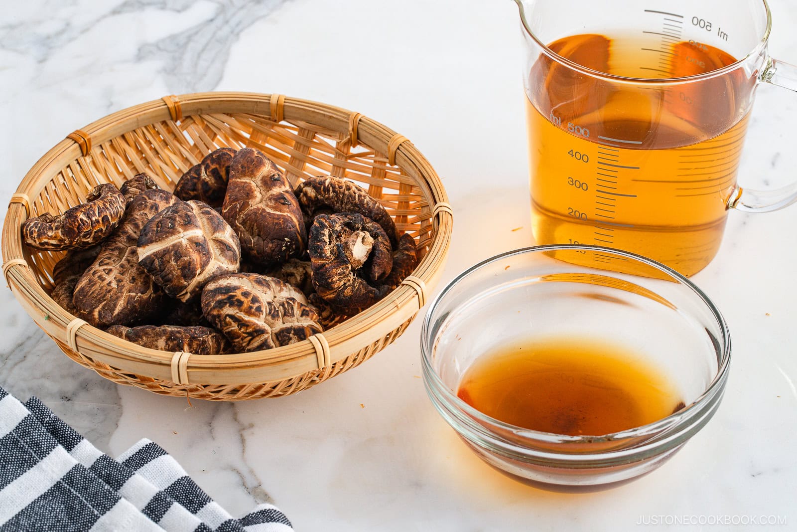 A basket of dried shiitake mushrooms, a glass measuring cup with light brown broth, and a small bowl of dark liquid on a marble countertop next to a striped cloth.