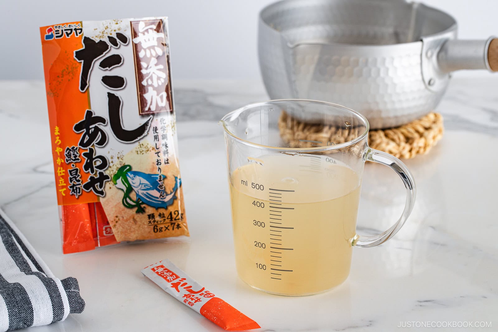 A clear measuring cup with light yellow dashi broth sits on a countertop next to a packet of dashi powder, an opened sachet, a pot, and a striped kitchen towel.