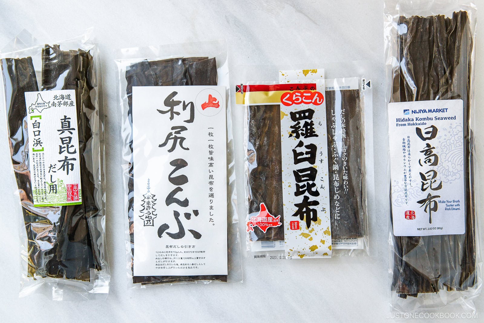 Four packages of dried kombu seaweed are arranged side by side on a white surface, each with distinct Japanese labels and clear packaging showing the dark, flat seaweed inside.
