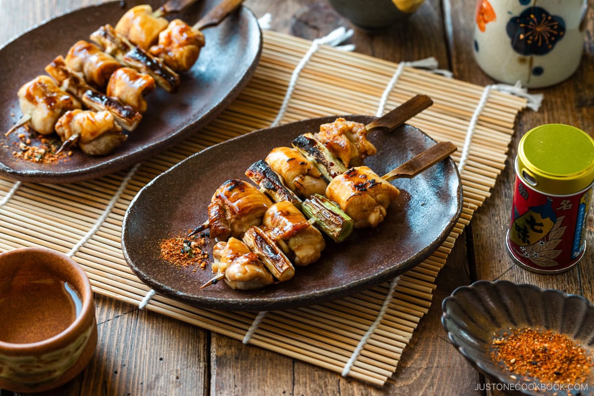 Oval rustic plates containing yakitori, Japanese grilled chicken and scallion skewers, coated with savory yakitori sauce and served with shichimi togarashi on the side.