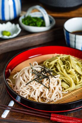 Zaru soba and zaru cha soba (cold buckwheat noodles and green tea noodles) served on a bamboo mat with shredded nori on top. Dipping sauce, green onions, and wasabi are in the background.